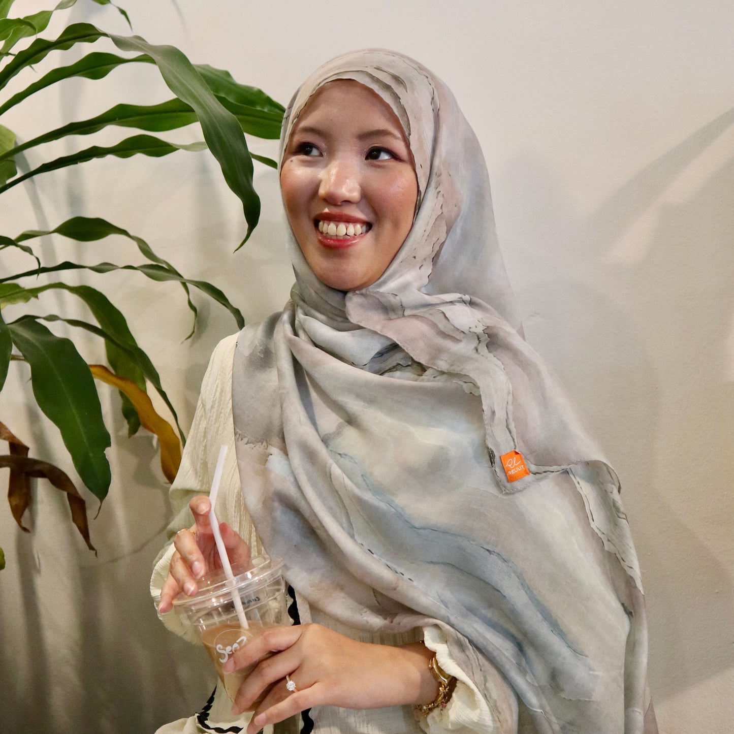 Woman wearing a light-colored hijab holding a drink with a straw, standing next to a plant.